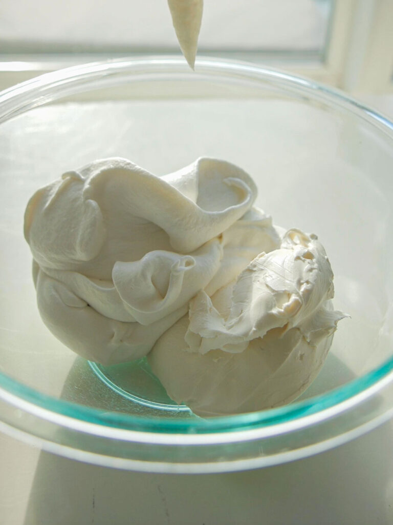 A glass bowl filled with smooth, creamy white frosting, with a dollop being piped in from above. The setting is well-lit, creating a soft, inviting appearance.